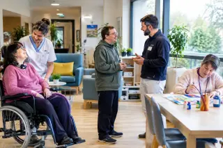 Salon lumineux d’un établissement médico-social avec résidents en situation de handicap, aide-soignant et éducateur, atmosphère accueillante et équipements adaptés.